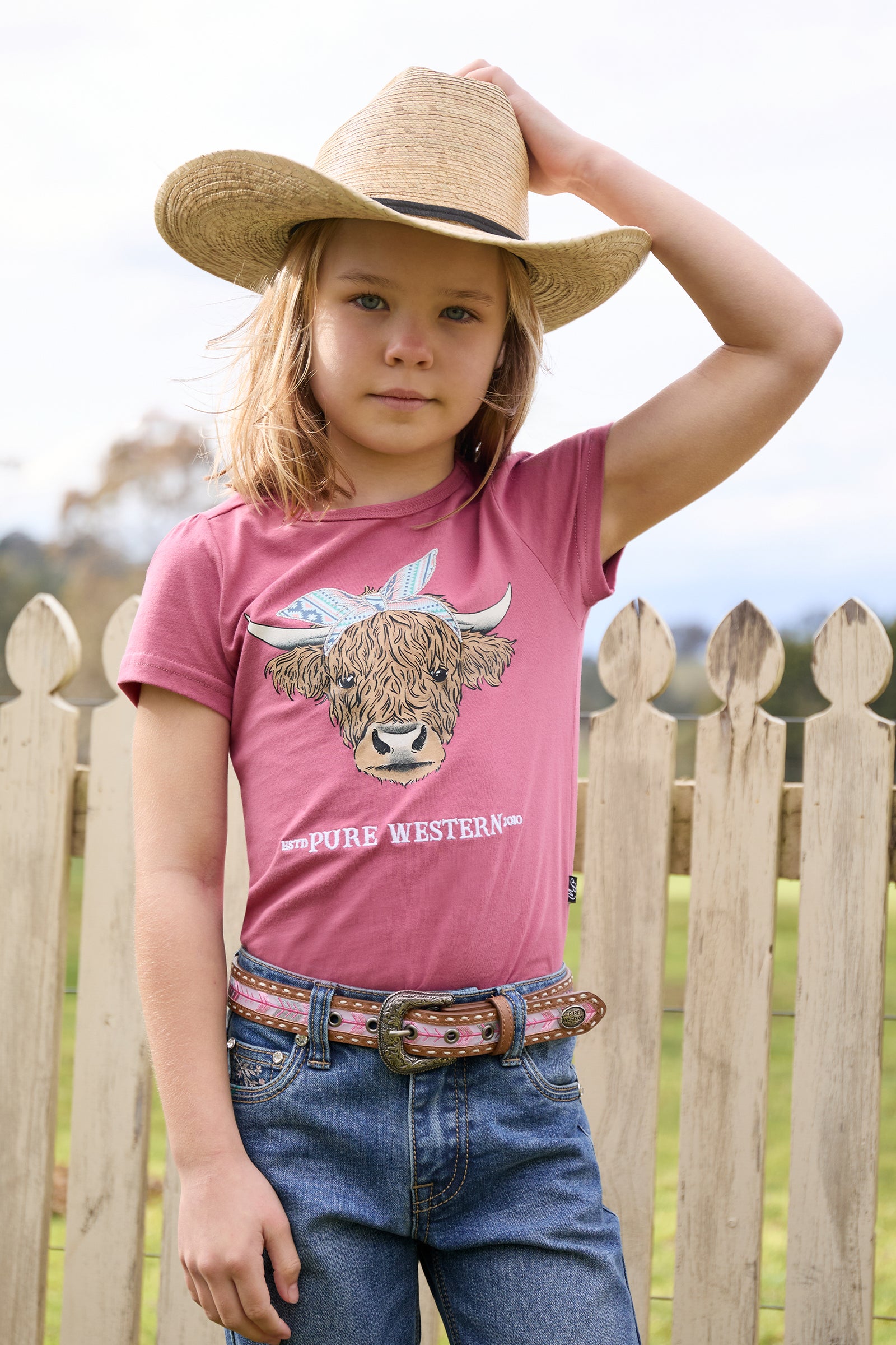 Child wearing a pink 'Pure Western' t-shirt with a cow graphic, blue jeans, and a straw hat, standing in front of a wooden fence.