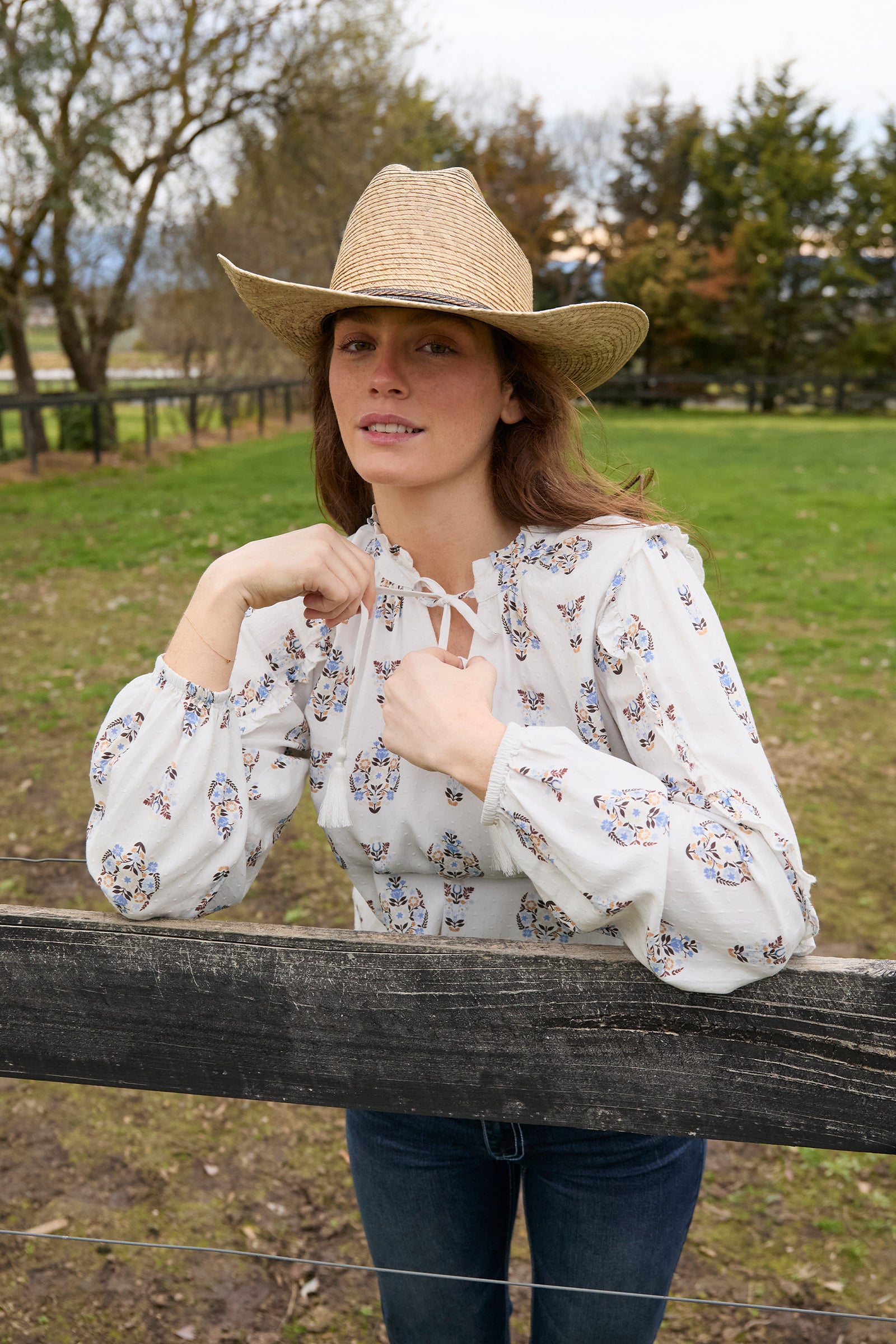 Woman wearing a straw hat and floral blouse standing by a wooden fence in an outdoor setting.