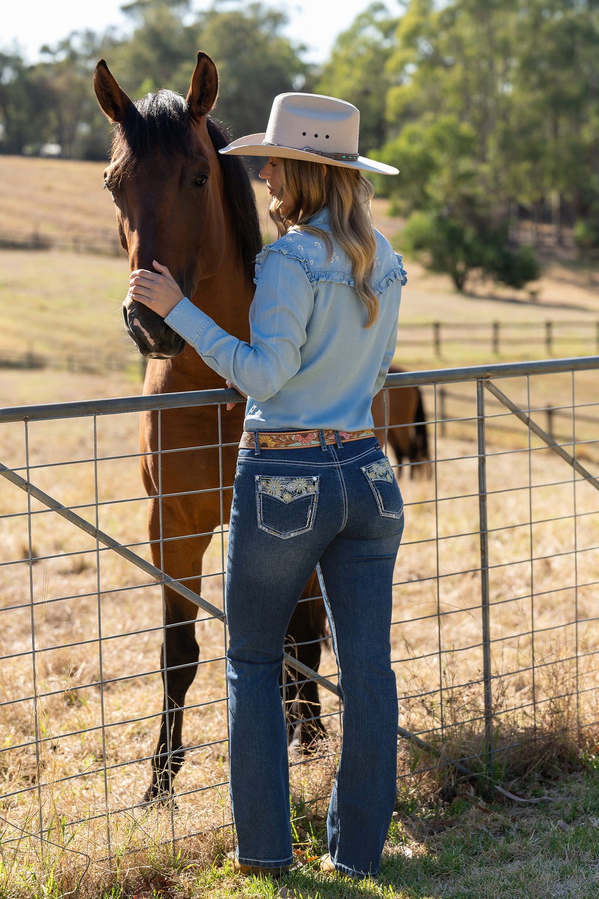 Pure Western Women's Jay Boot Cut Jean. Woman in cowboy hat and light blue shirt standing next to a horse behind a fence in a rural setting.