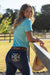 Pure Western Women's Ana Boot Cut Jean. Woman in a turquoise shirt and jeans standing by a fence with a blurred background