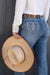 Pure Western Women's Pam High Rise Boot Cut Jean. Person wearing blue jeans with a decorative pocket, holding a straw hat against a dark background