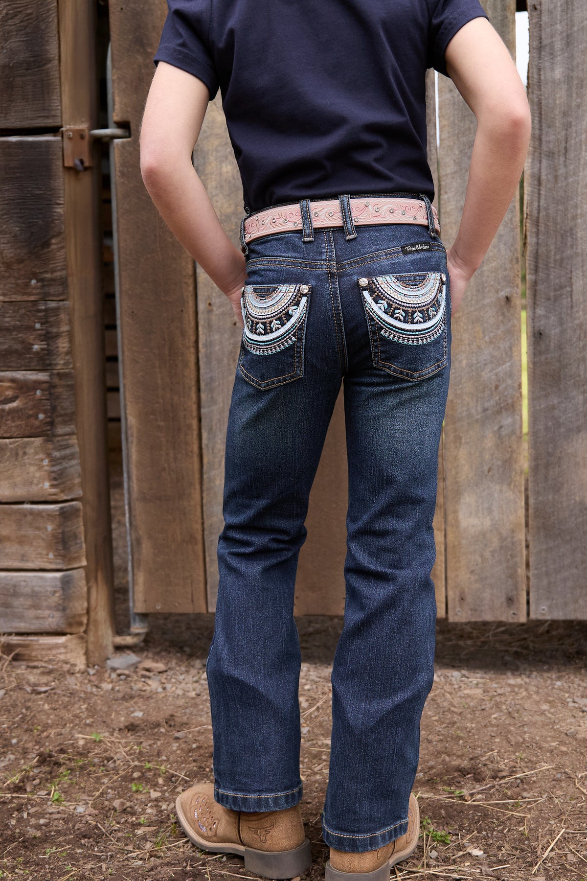Person wearing blue jeans with decorative back pockets in front of a wooden fence.