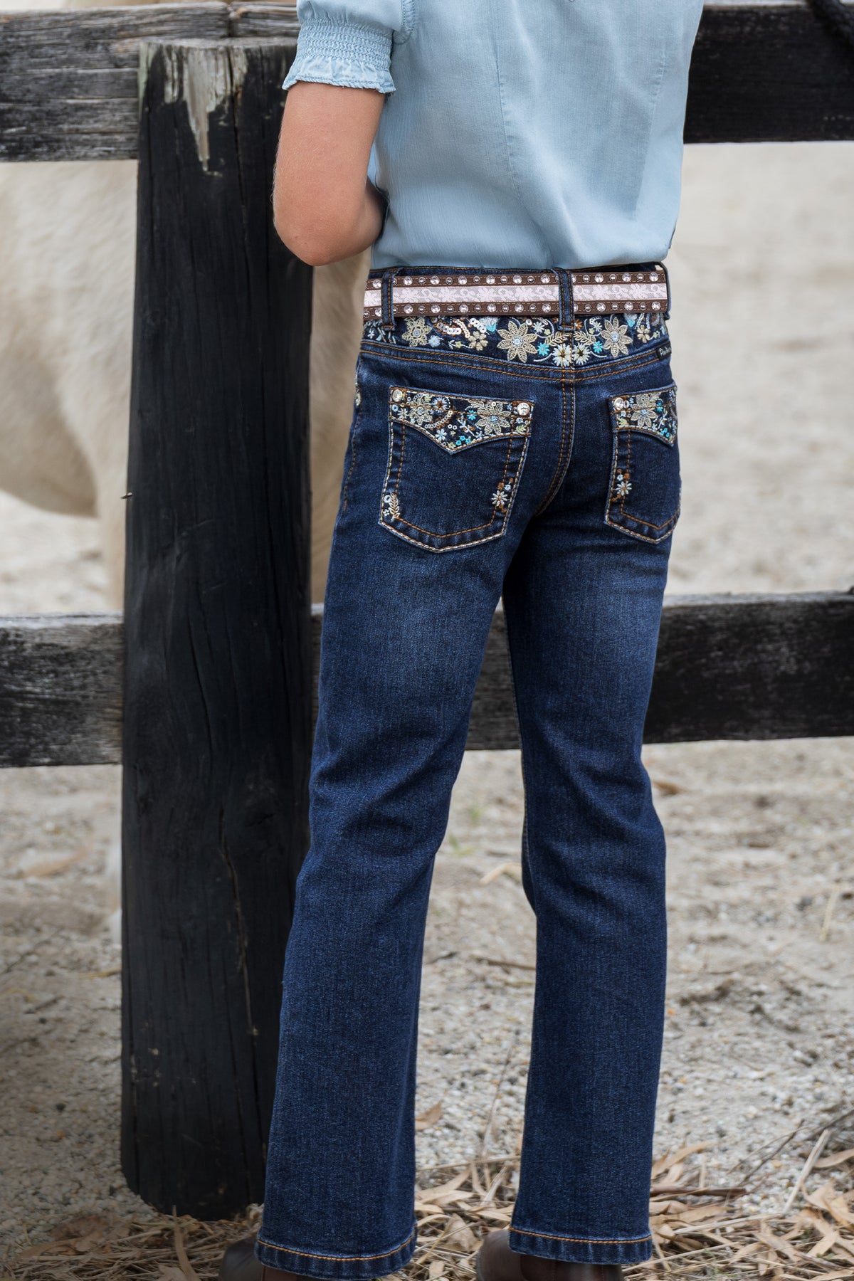 A child wearing dark blue denim jeans with decorative floral embroidery on the back pockets and a patterned belt, standing outdoors next to a wooden fence.