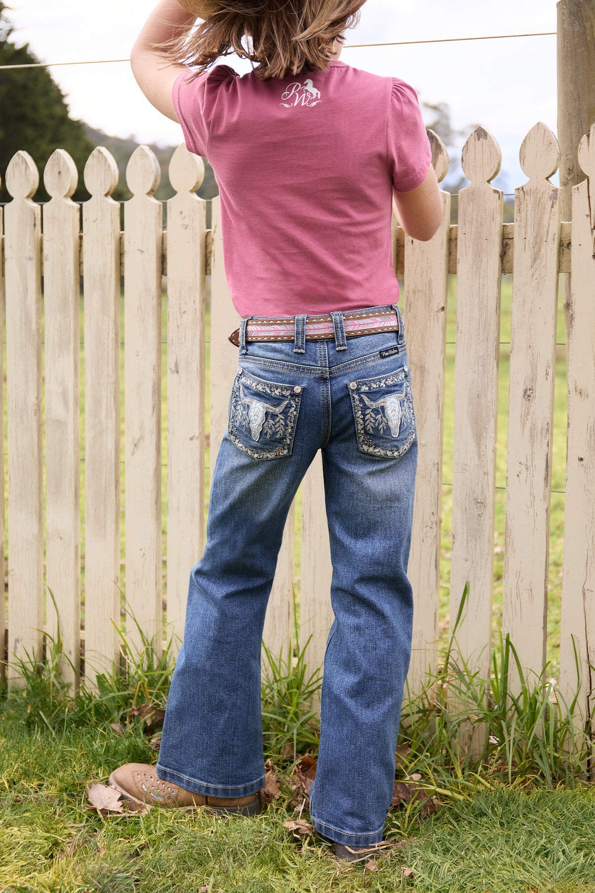 Person wearing a pink shirt and blue jeans standing in front of a wooden fence.