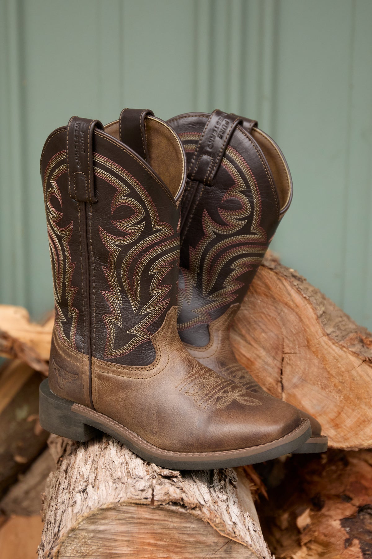 Brown cowboy boots on a stack of wooden logs against a green metal background