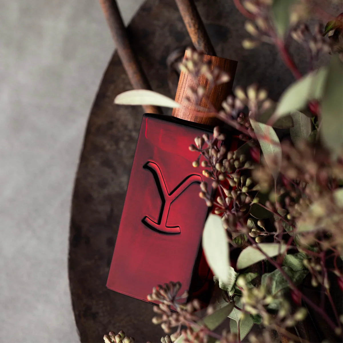 A rectangular dark red glass bottle with a carved red symbol on the front, set on a rustic metal surface surrounded by dried flowers and greenery.