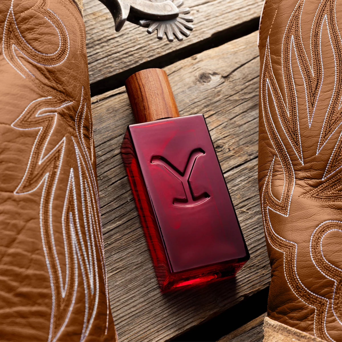 A rectangular bottle of red liquid with a wooden cap and an embossed Y-shaped logo on the front, placed on a rustic wooden surface between two brown leather cowboy boots with white stitching.