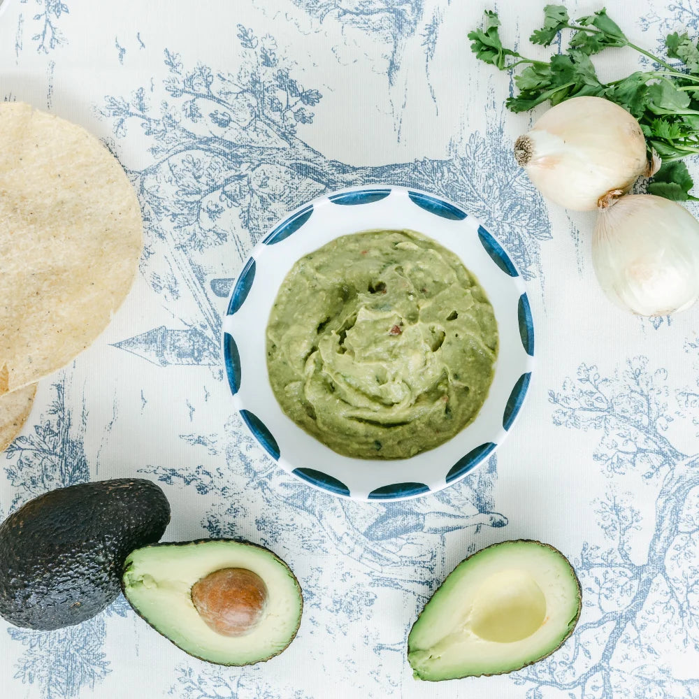 Avocado dip in a bowl surrounded by avocados, onions, and cilantro on a decorative surface.