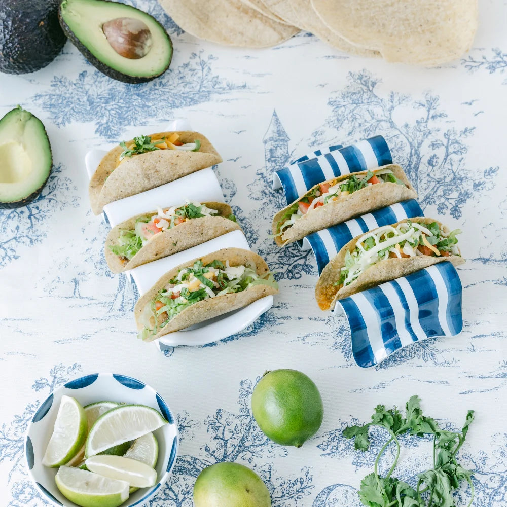 Small tacos on a decorative plate with avocados, limes, and tortillas on a patterned surface.