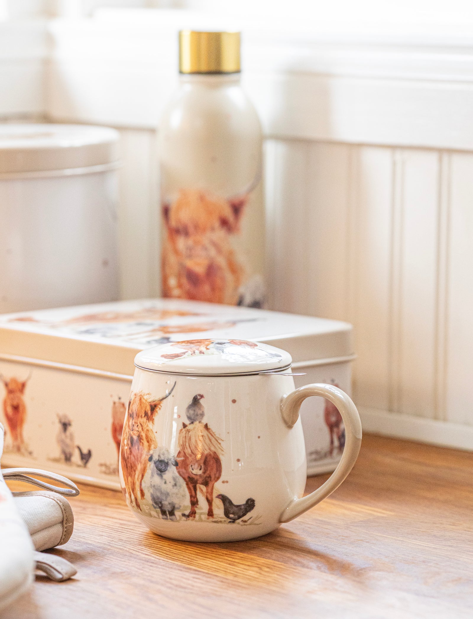 A ceramic mug with a lid, decorated with illustrations of dogs and chickens, sits on a wooden table in a bright, sunlit room. A matching tin box and a water bottle with a dog print are visible in the background.