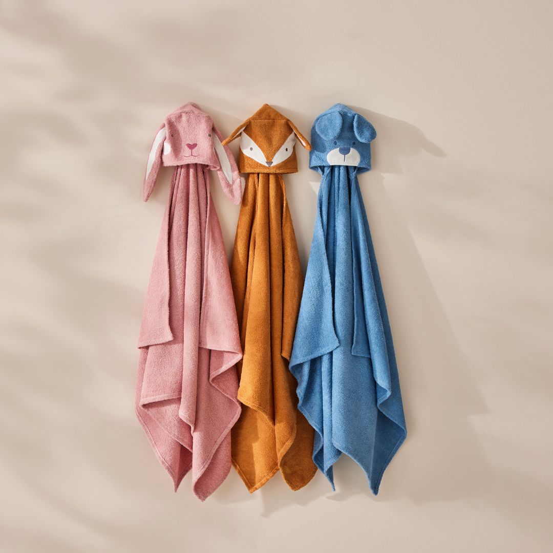 Three hooded towels in pink, orange, and blue hanging against a beige background