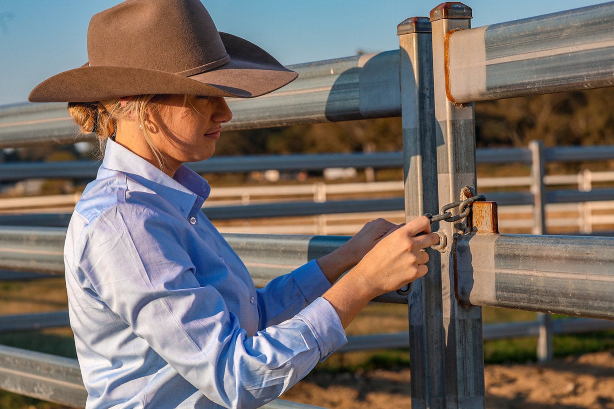 Reba Women's Blue Bird Oxford Gal L/S Shirt. Two women wearing cowboy hats in a rural setting with a fence and trees in the background.
