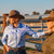 Reba Women's Blue Bird Oxford Gal L/S Shirt. Two women wearing cowboy hats in a rural setting with a fence and trees in the background.