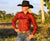 Reba Women's Maroon Linen Gal L/S Shirt. A woman wearing a maroon long-sleeve shirt and a white cowboy hat, sitting by a body of water.
