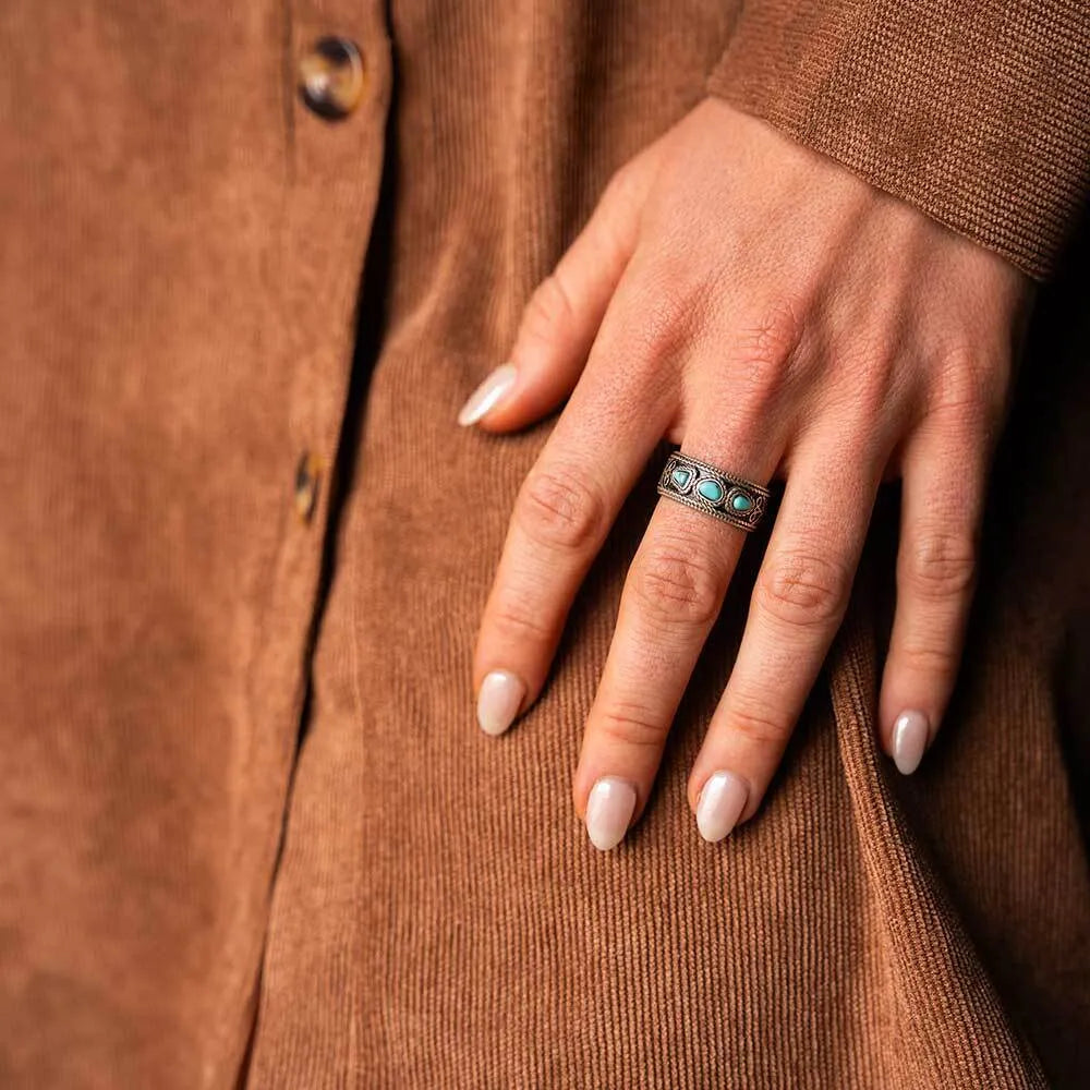 A close-up of a hand wearing a silver ring with three small turquoise-blue stones, resting on a brown corduroy jacket.