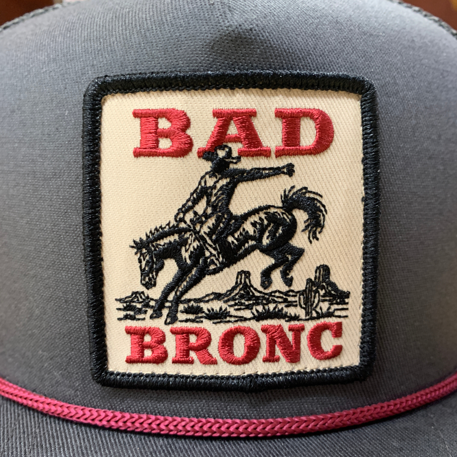 A close-up of a gray trucker hat featuring a square embroidered patch on the front. The patch has a cream background with a black border and shows a cowboy riding a bucking bronco, with the words 'BAD BRONC' embroidered in red.
