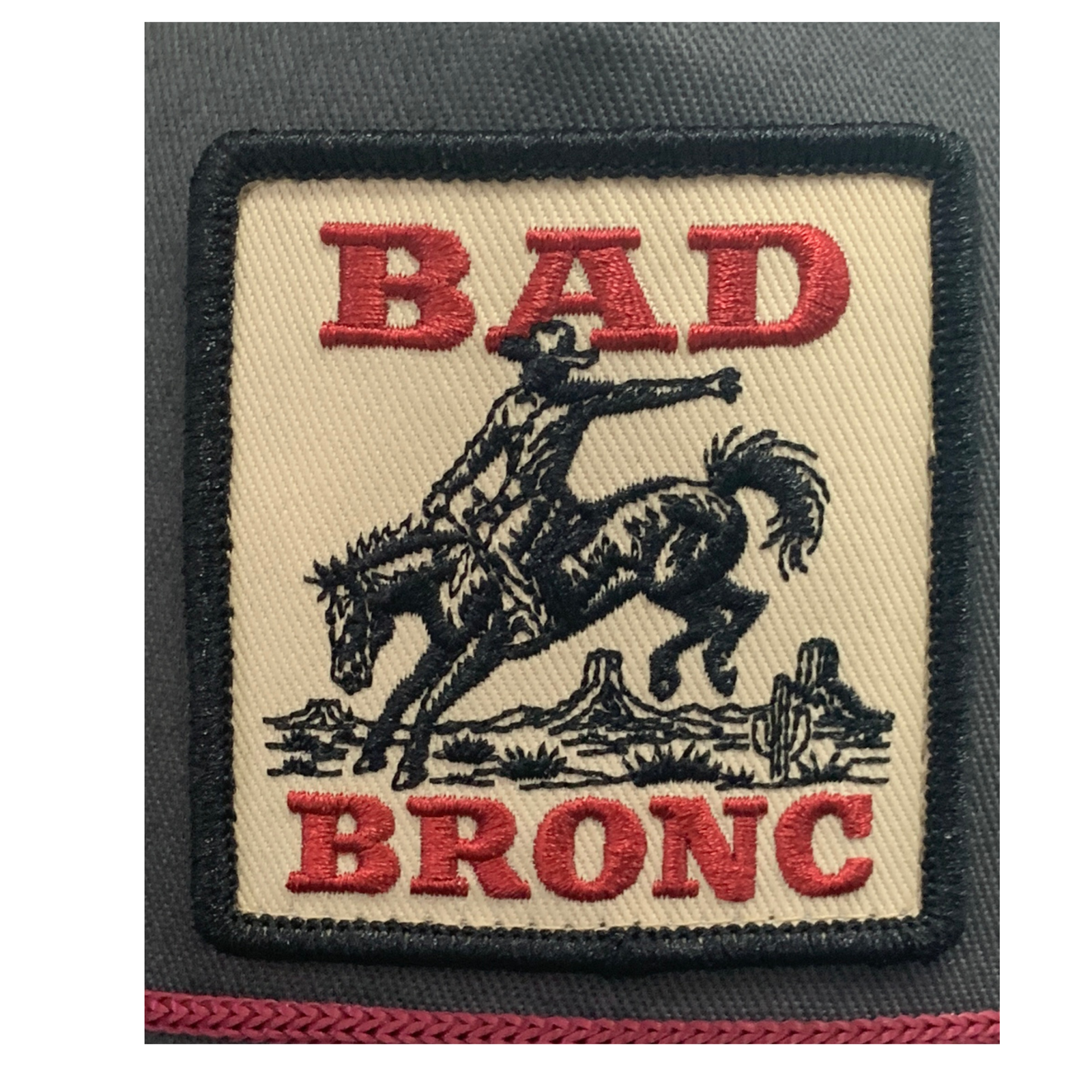 An embroidered rectangular patch with a black border on a dark gray fabric background. The patch has a beige center with red embroidered text reading 'BAD BRONC' and a black cowboy riding a bucking horse, with a cactus and mountains below.