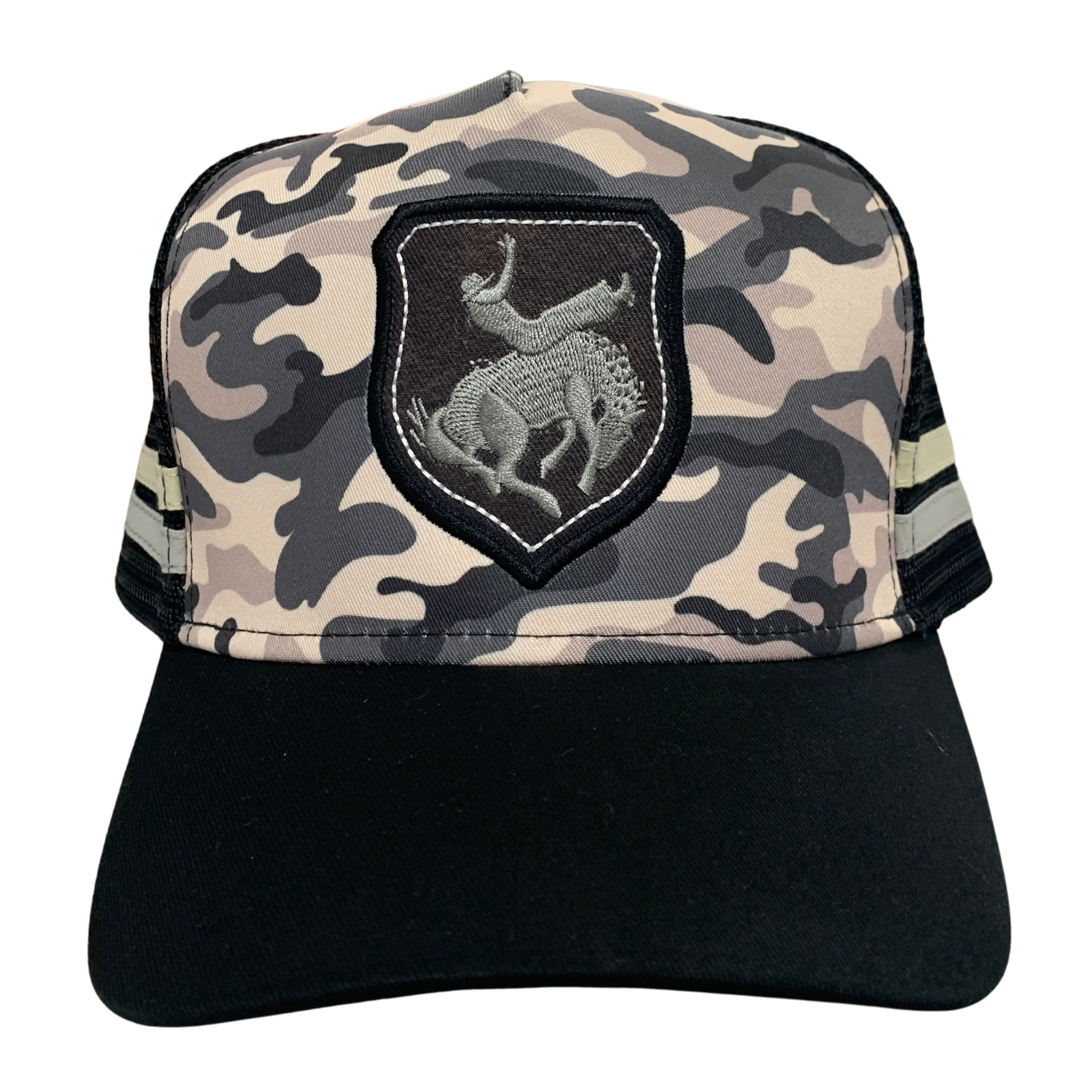 A trucker-style baseball cap with a black curved brim, black mesh back, and a front panel featuring a beige, gray, and black camouflage pattern. A black shield patch with a silver-toned logo is stitched on the left front.