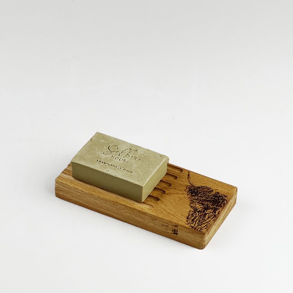A rectangular bar of olive green soap resting on a carved wooden soap dish with a dark engraved design, photographed on a clean white background.