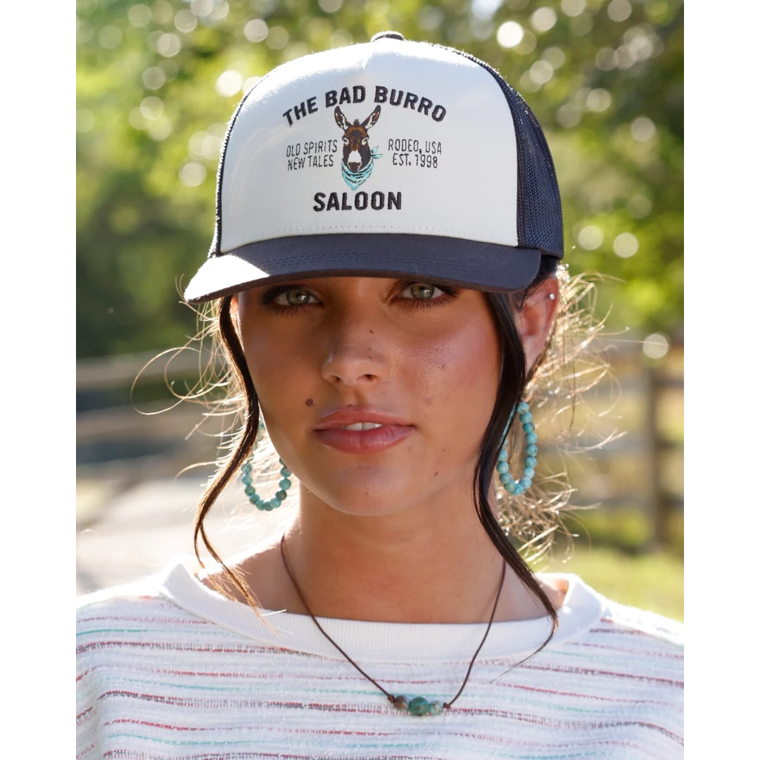 A woman wearing a white and dark green trucker hat with 'THE BAD BURRO SALOON' printed on the front, set against a blurred outdoor background.