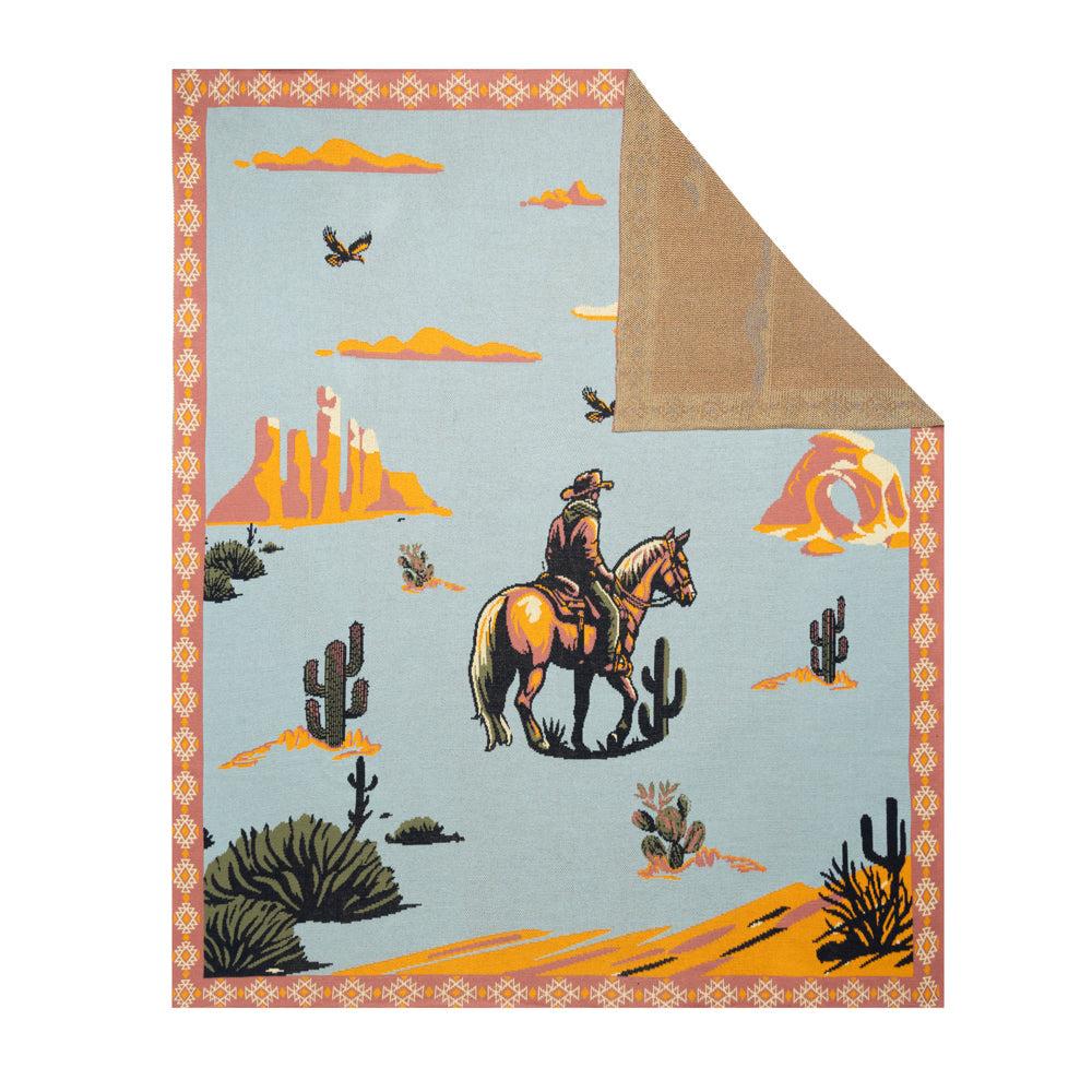 Lone Bronco Rider Throw. A throw blanket featuring an illustration of a cowboy riding a bronco with a desert landscape, cacti, and mesas on a blue background. The blanket is bordered with a tribal-inspired pattern and has a brown backside.