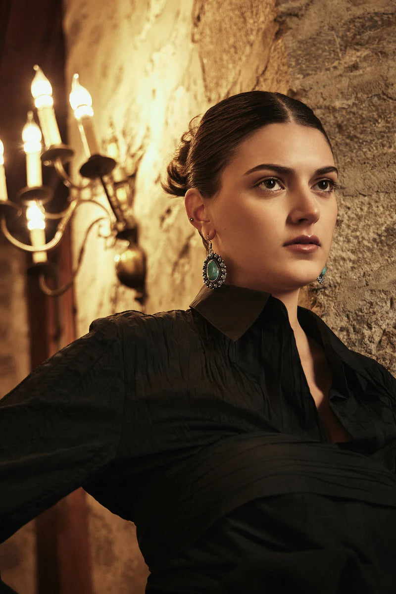 Woman in a black outfit with earrings standing against a stone wall with a chandelier.