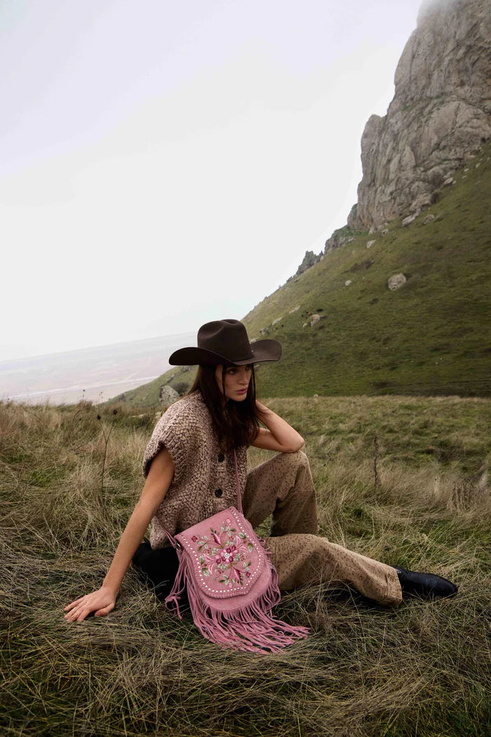 Person sitting in a grassy field with a pink bag, wearing a cowboy hat and patterned shirt.