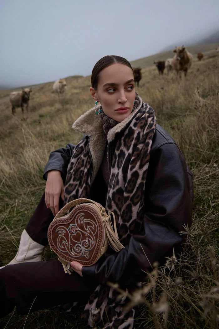 Woman in a field with cows, wearing a leopard print scarf and holding a woven bag.
