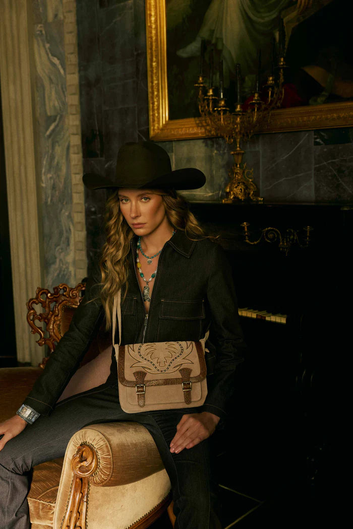Woman in a black outfit with a cowboy hat and a beige bag, sitting in an ornate room.