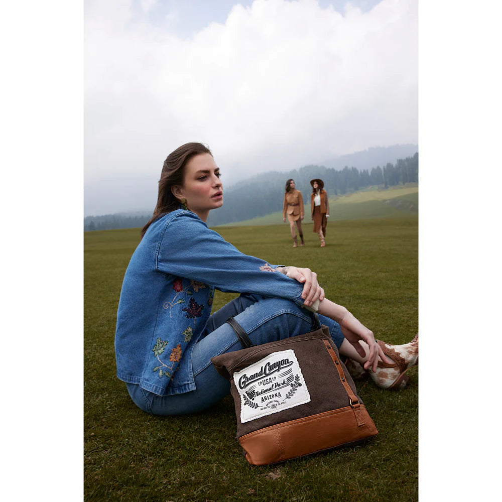 Woman sitting on grass with a brown leather bag, with two people in the background.