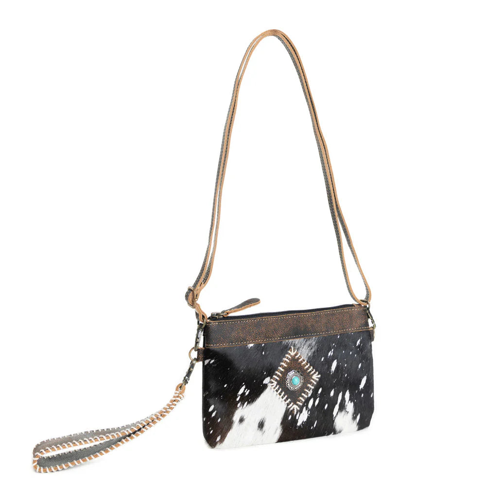 Eye of the Goddess Crossbody Bag in Black. Black and white cowhide handbag with a decorative clasp on a white background