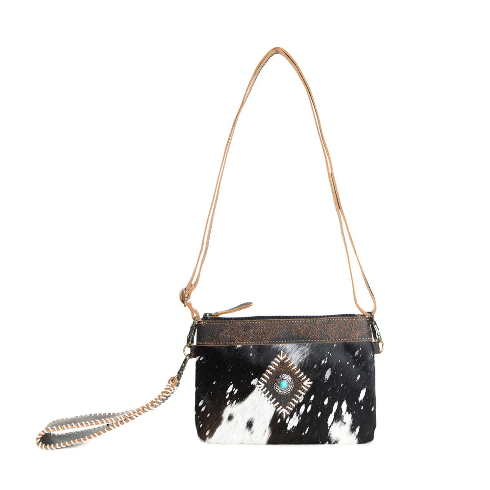 Eye of the Goddess Crossbody Bag in Black. Black and white cowhide handbag with gold chain and decorative element on a white background