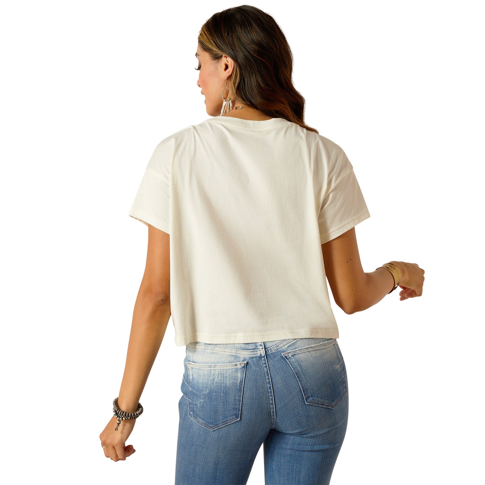 Ariat Women's Wild Hat Cropped S/S T-Shirt. A woman wearing a beige, solid color, cropped T-shirt with a boxy fit, paired with blue denim jeans. She has accessorized with a long necklace and multiple bracelets.