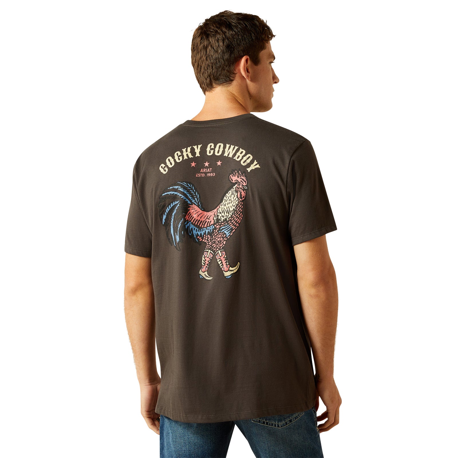 Ariat Men's Cowboy Cocky T-Shirt. Man wearing a dark brown t-shirt with a colorful rooster graphic and 'Cocky Cowboy' text on the back.