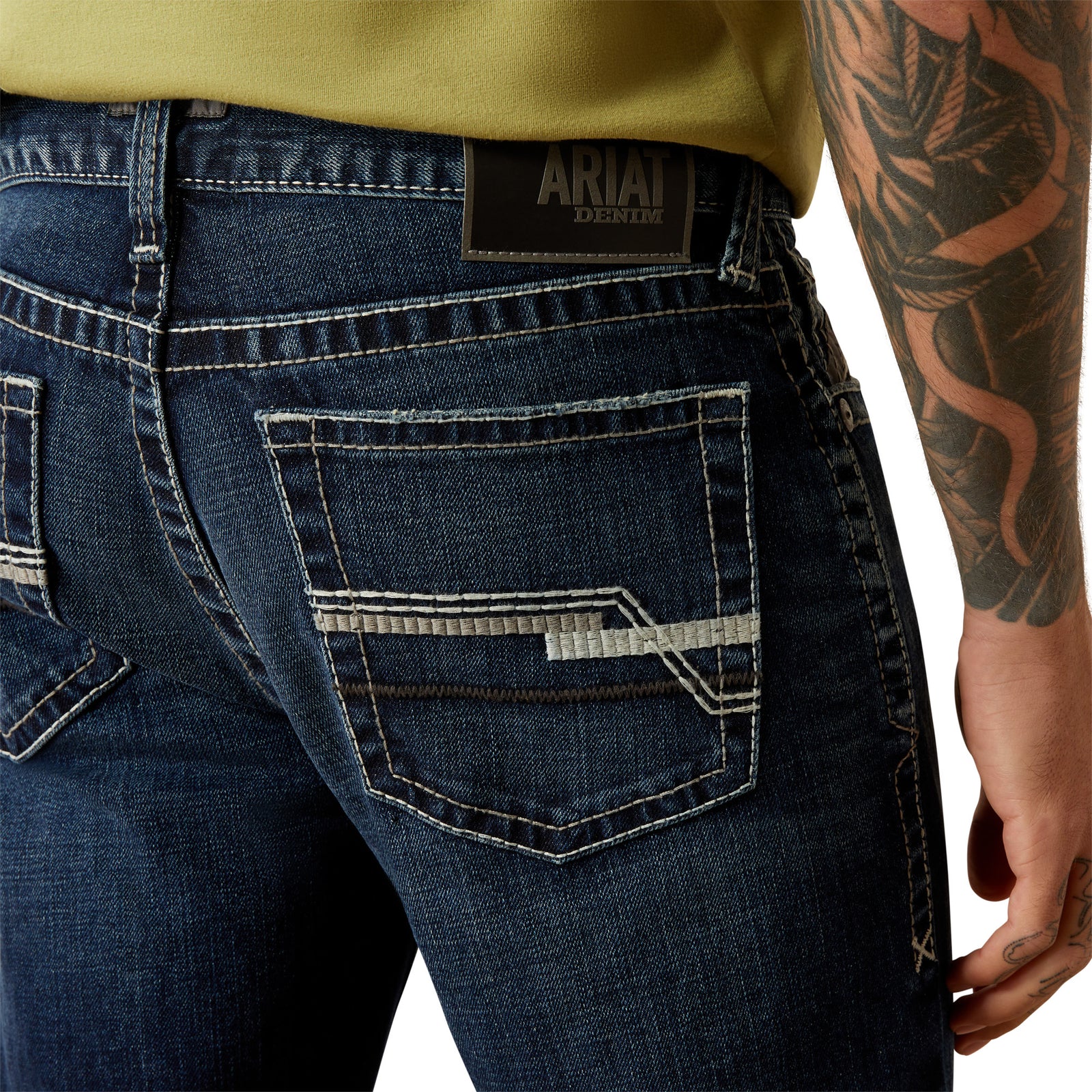 Ariat Men's M8 Modern Matthew Straight Leg Jean. Close-up of blue jeans with a tattooed arm and Ariat logo.