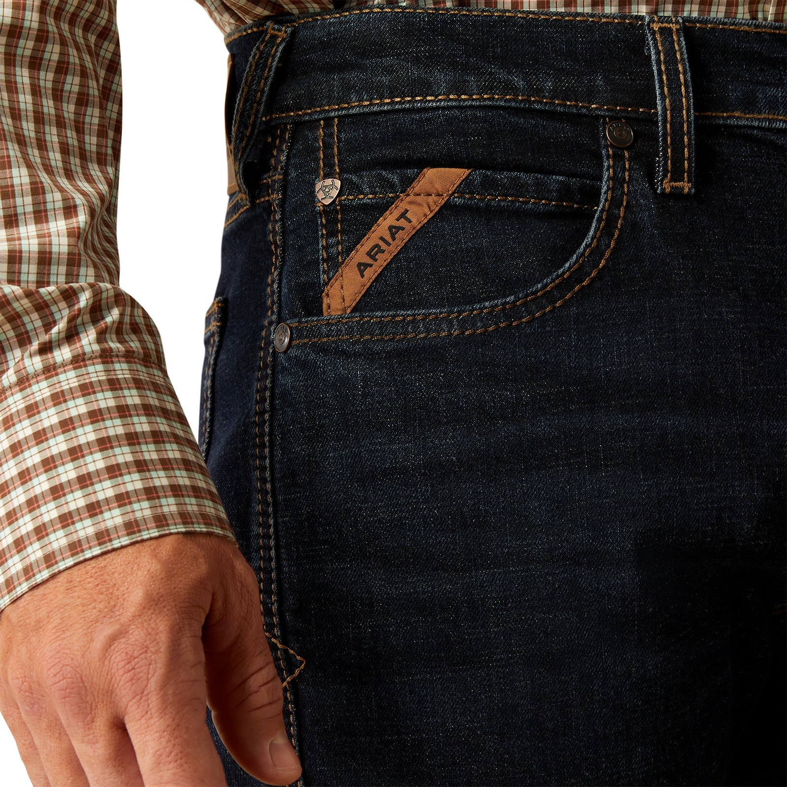 Ariat Men's M5 Dexter Straight Leg Jean. Close-up of dark blue jeans with Ariat logo on pocket, worn by a person with plaid shirt sleeve.