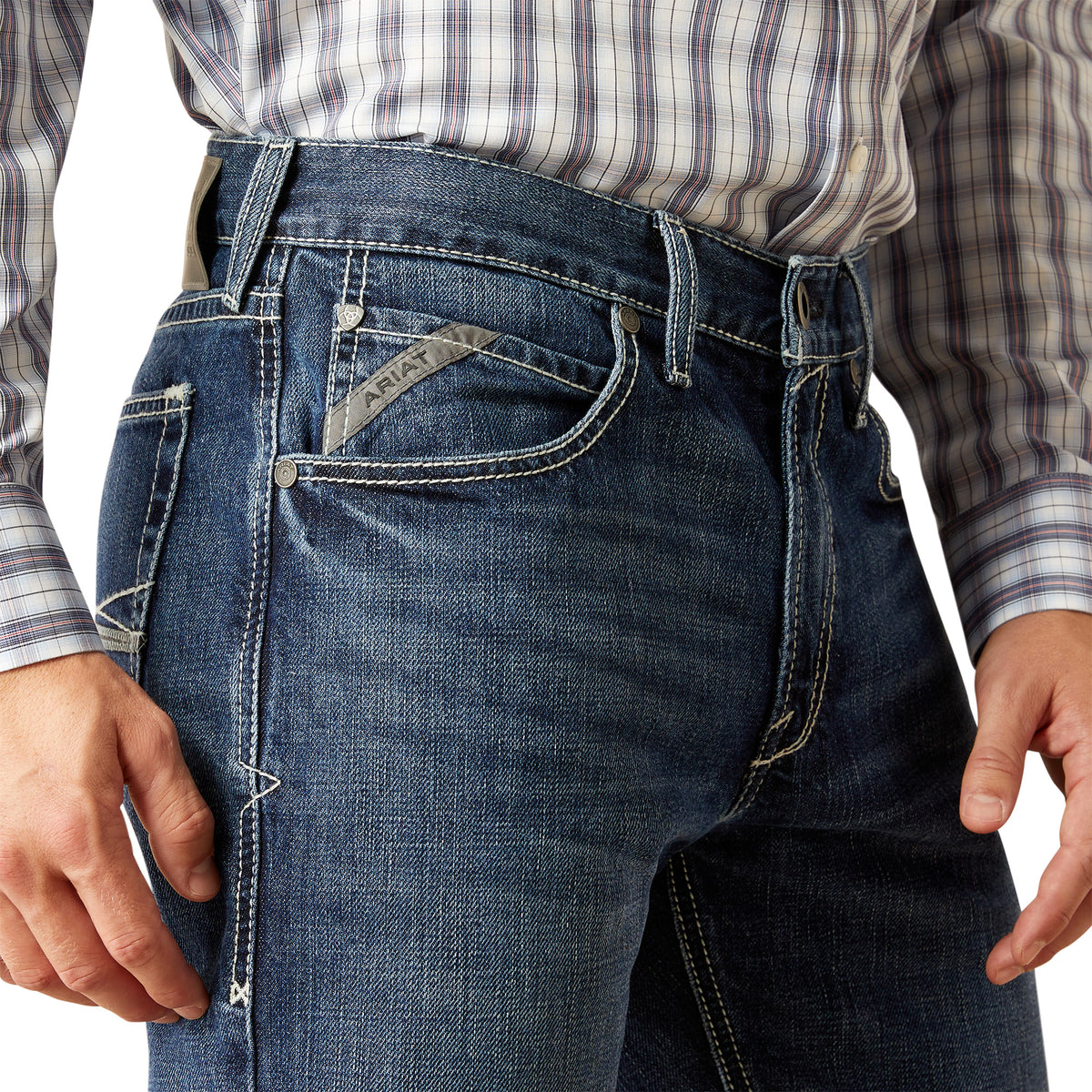 Ariat Men's M2 Camilo Boot Cut Jean. Close-up of blue jeans with a plaid shirt worn underneath on a white background