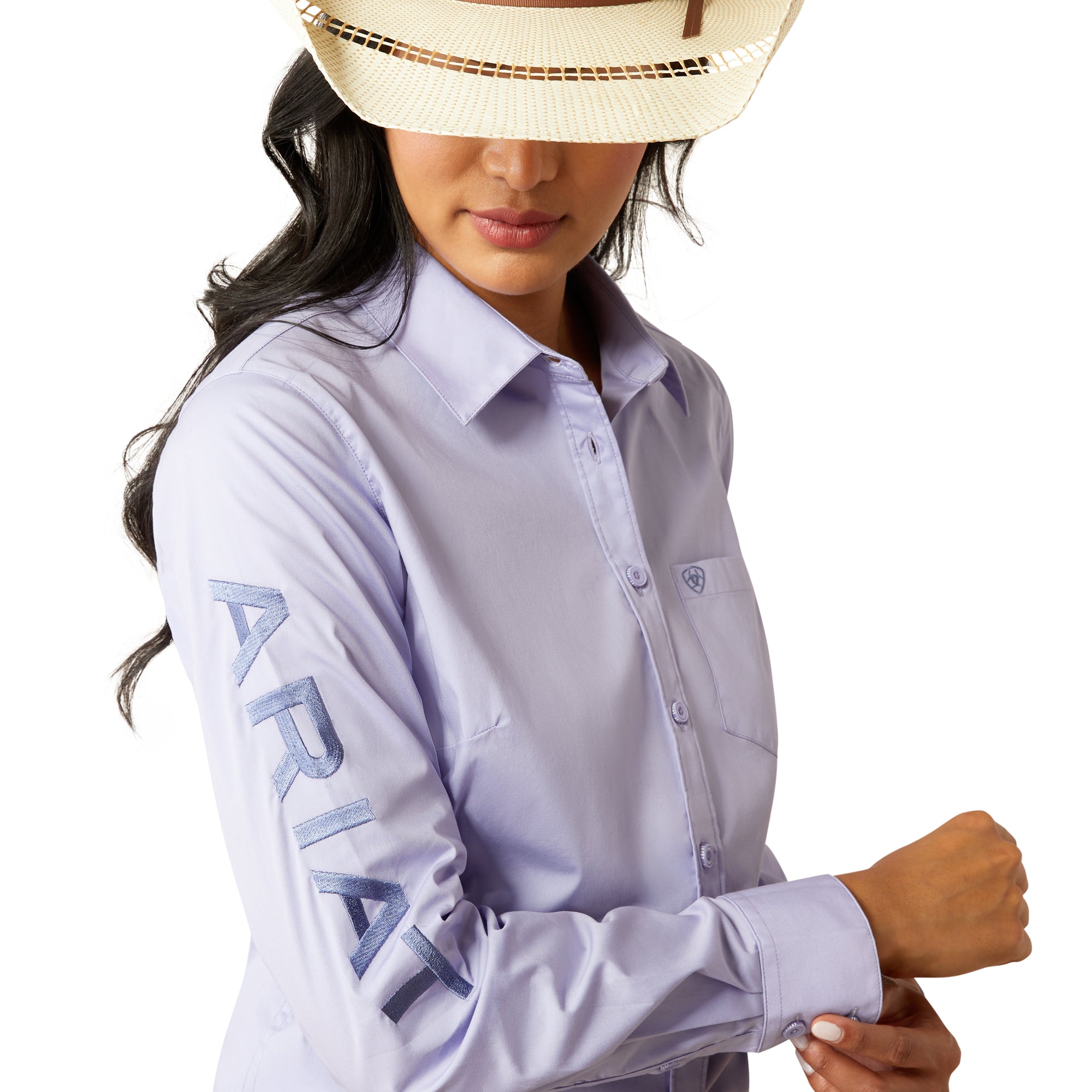Ariat Women's Wrinkle Resistant Team Kirby L/S Western Shirt. Woman wearing a light purple shirt, blue jeans, and a cowboy hat on a white background