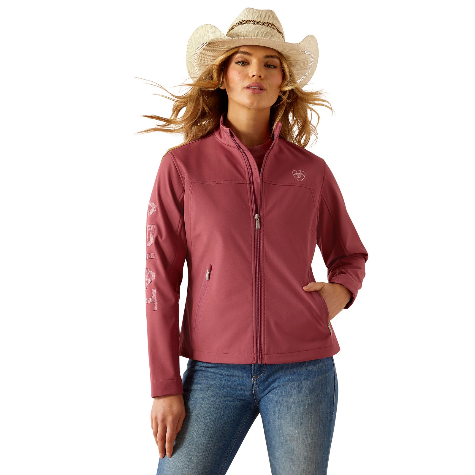 Ariat Women's Team Softshell Jacket. Woman wearing a pink jacket and blue jeans with a white background
