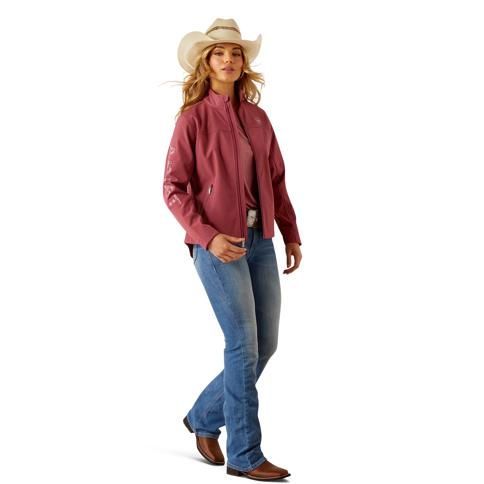 Ariat Women's Team Softshell Jacket. Woman wearing a pink jacket, blue jeans, and a cowboy hat on a white background