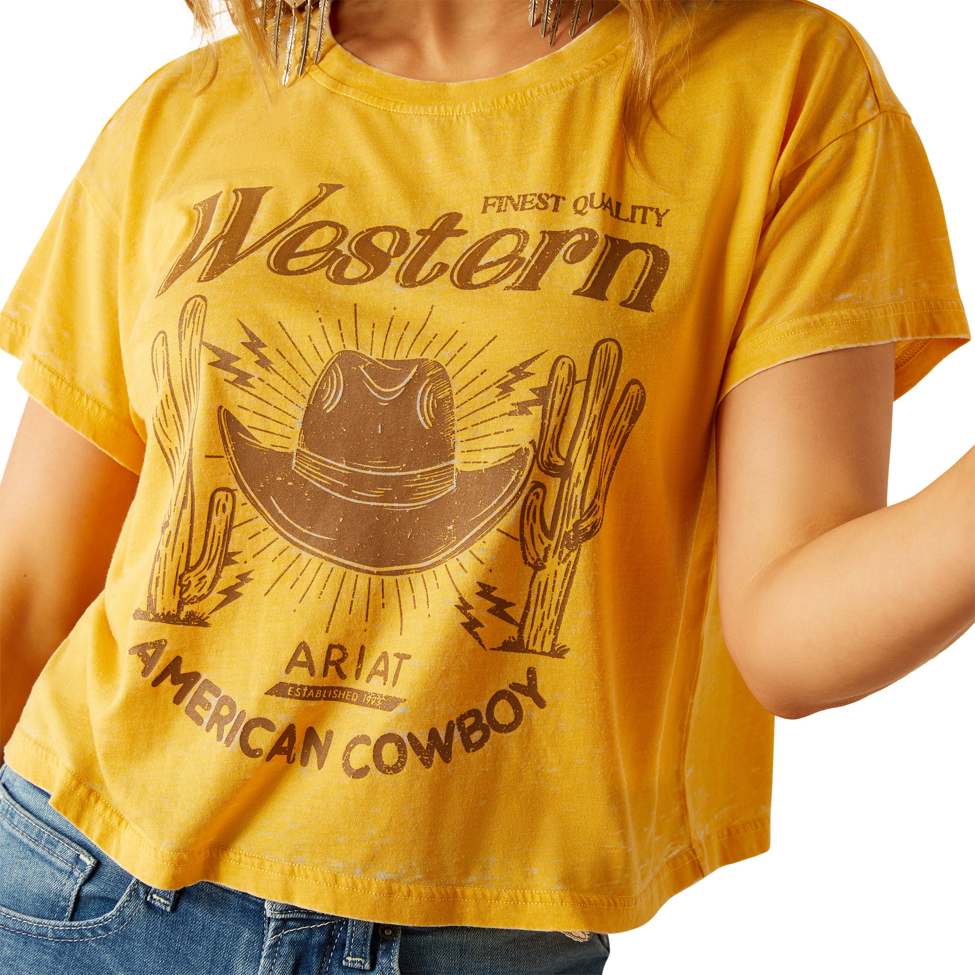 Ariat Women's Gold Rush Crop T-Shirt. Woman wearing a yellow 'Western' t-shirt and denim shorts on a white background