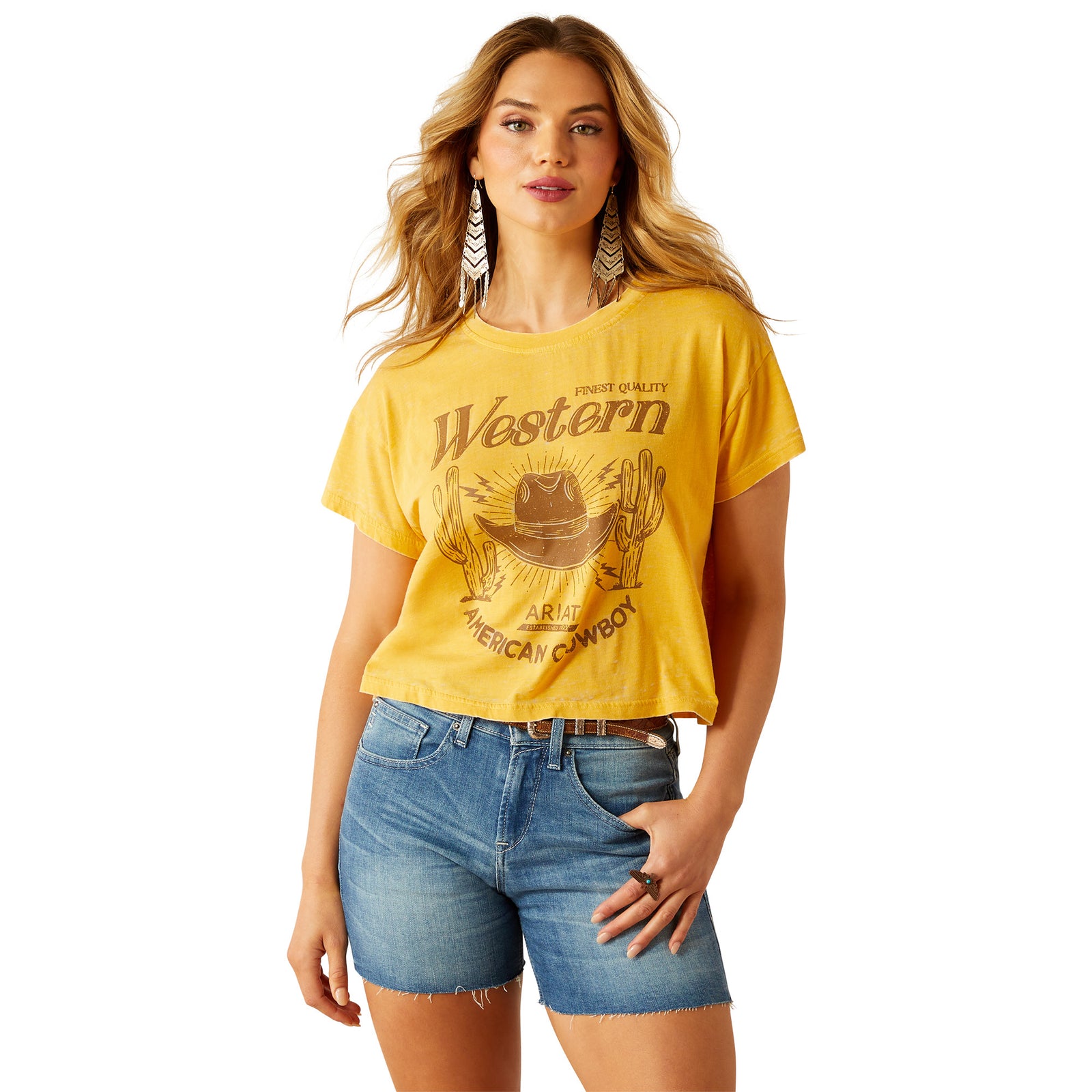 Ariat Women's Gold Rush Crop T-Shirt. Woman wearing a yellow 'Western' t-shirt and denim shorts on a white background