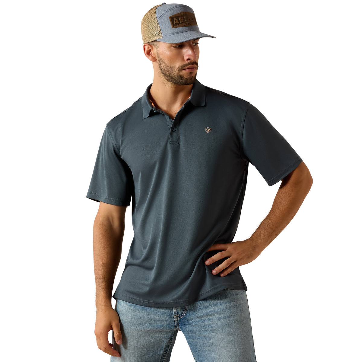A man wearing a dark slate blue short-sleeve polo shirt with a small embroidered shield logo on the chest, paired with a gray and tan trucker hat and light blue jeans, against a solid white background.
