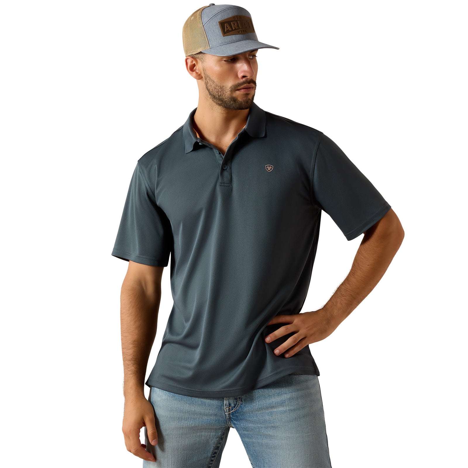 A man wearing a dark slate blue short-sleeve polo shirt with a small embroidered shield logo on the chest, paired with a gray and tan trucker hat and light blue jeans, against a solid white background.