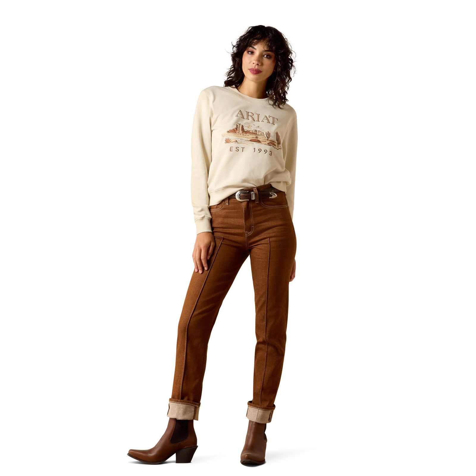 Woman wearing a cream sweater with a graphic design and brown pants on a white background