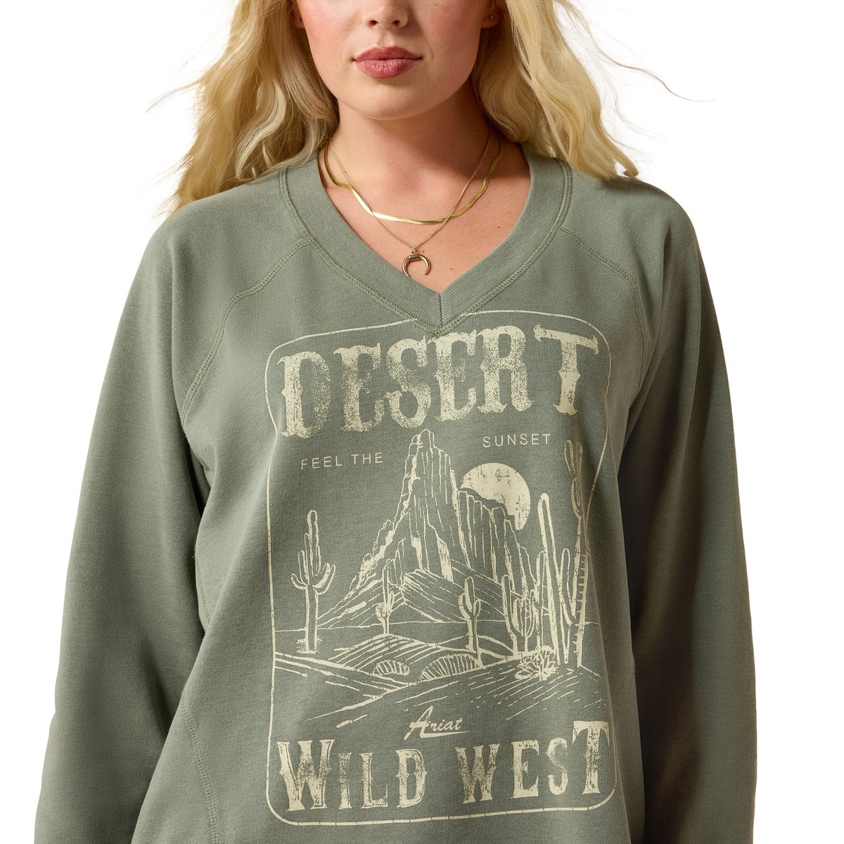 A woman wearing a sage green V-neck sweatshirt with a distressed desert graphic that reads 'DESERT WILD WEST' and 'FEEL THE SUNSET'.