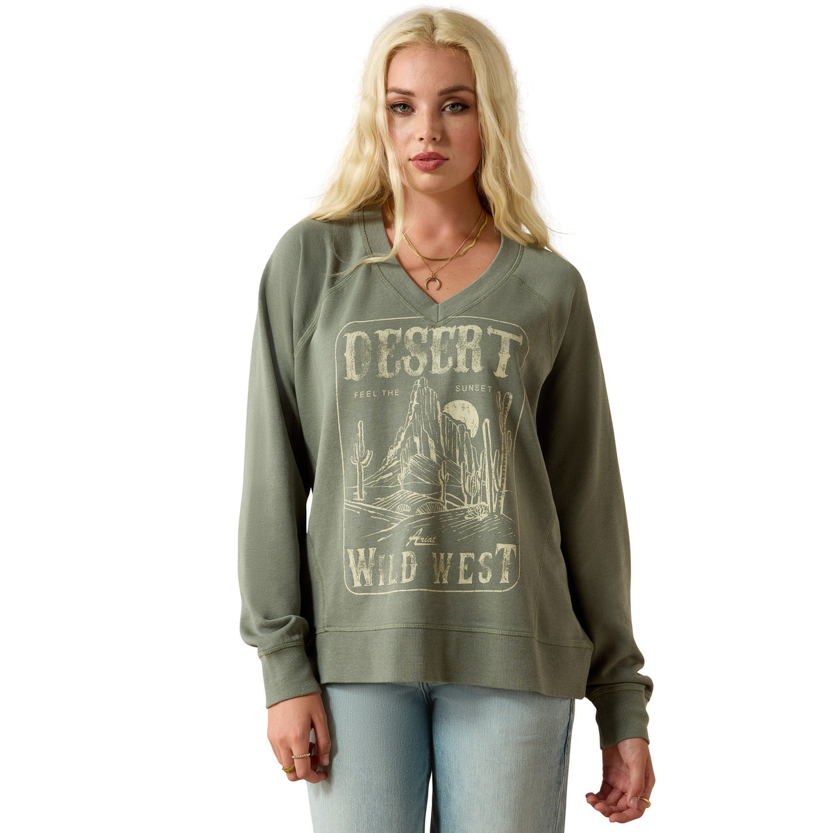 A blonde woman modeling an olive green long-sleeve V-neck sweatshirt with a distressed beige 'Desert Wild West' graphic on the front, paired with light blue jeans against a plain white background.