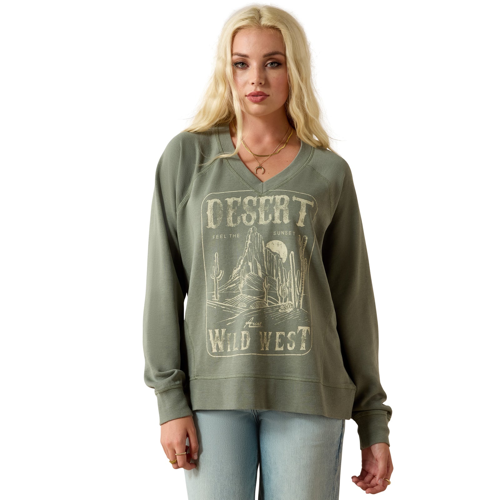 A blonde woman modeling an olive green long-sleeve V-neck sweatshirt with a distressed beige 'Desert Wild West' graphic on the front, paired with light blue jeans against a plain white background.
