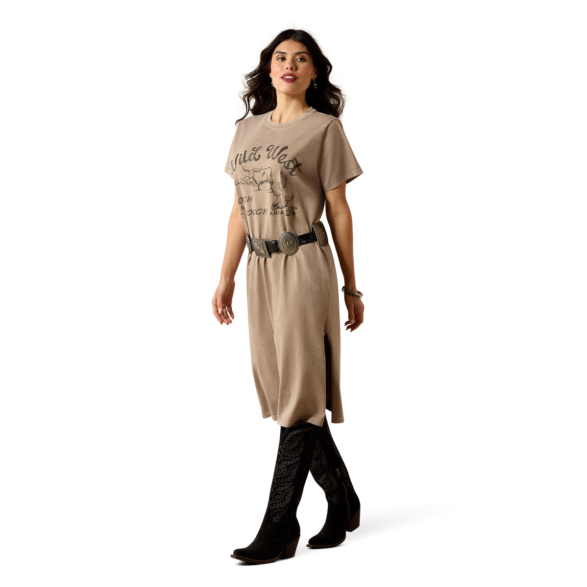 A woman modeling a beige graphic t-shirt dress with a western print, styled with a black belt, silver conchos, and black cowboy boots against a white studio background.