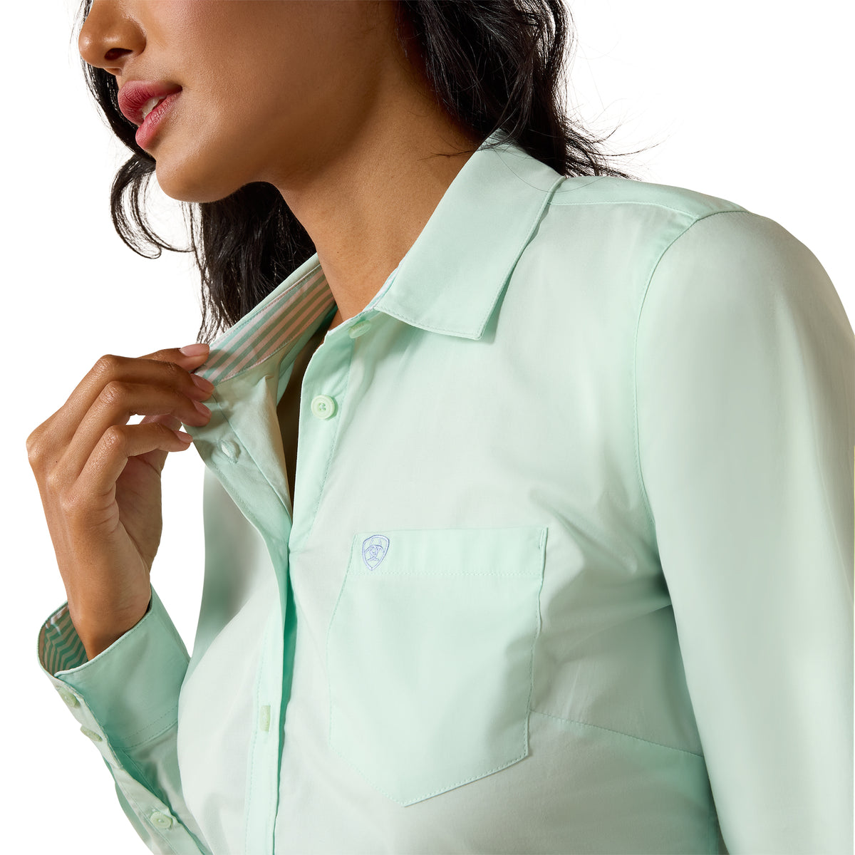 A close-up of a woman wearing a light mint green long-sleeve button-up shirt, adjusting the collar with one hand. The shirt features a classic pointed collar, a chest pocket with a small embroidered logo, and a clean white background.